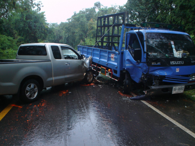 Ein schwerbeschädiger Lkw mit eingedrückter Front und eingebeulter Karosserie liegt am Straßenrand, umgeben von Bäumen unter einem klaren blauen Himmel.