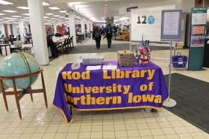 Eine Ausstellung mit einem Tisch, auf dem verschiedene Landesflaggen, eine Box, Papiere und ein Banner liegen, eine Kugel links daneben, ein Anschlagbrett rechts daneben und mehrere Tische und Stühle links.