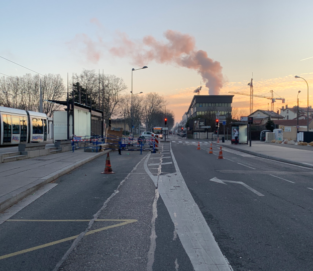 Eine Straßenansicht mit Verkehrskegeln, einem Bus, Laternenpfählen, Bäumen, Gebäuden, Kränen und Rauch, der aus einem fernen Gebäude aufsteigt.