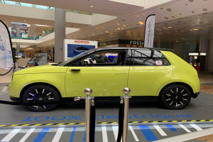 Ein gelbes wasserstoffbetriebenes Elektroauto steht drinnen auf einem Parkplatz, umgeben von Absperrpollern mit Seilen, Bannern und Schildern, mit einer Person im Auto und einer Gruppe von Menschen in der N"he.