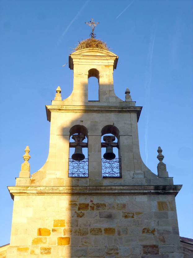 Ein Kirchturm mit einem Kreuz darauf, umgeben von einem Gebäude und einem klaren blauen Himmel, geschmückt mit Glocken und einem Geländer.