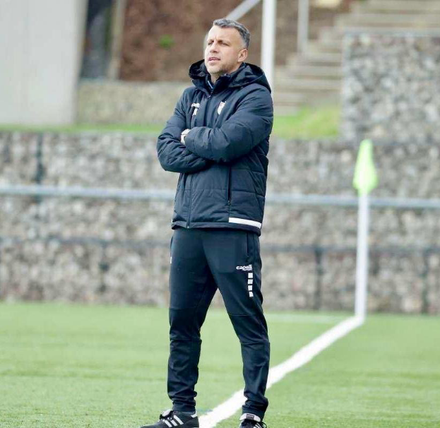 Ein Mann steht auf einem Fußballfeld mit verschränkten Armen, trägt eine schwarze Jacke und Hose und hat im Hintergrund einen Zaun, Treppe und Wand.