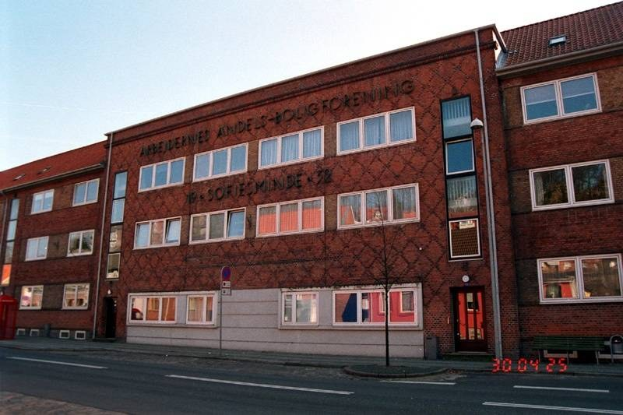 Ein großes Backsteingebäude mit vielen Fenstern, das als Hauptquartier der deutschen Bundespolizei dient, das auf einer Straße mit einem Gehweg, einem Baum, einer Bank, einem Laternenmast und einer roten Box steht, mit dem Himmel im Hintergrund.