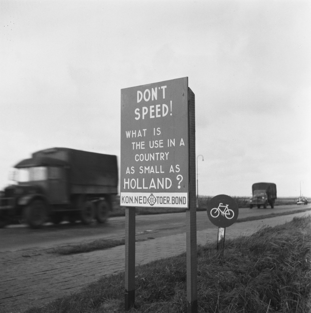 Schwarz-weißes Foto eines Lkws auf einer Straße mit einem "Fahren Sie nicht zu schnell"-Schild im Vordergrund und anderen Fahrzeugen, Gras, Pfosten und einem klaren Himmel im Hintergrund.