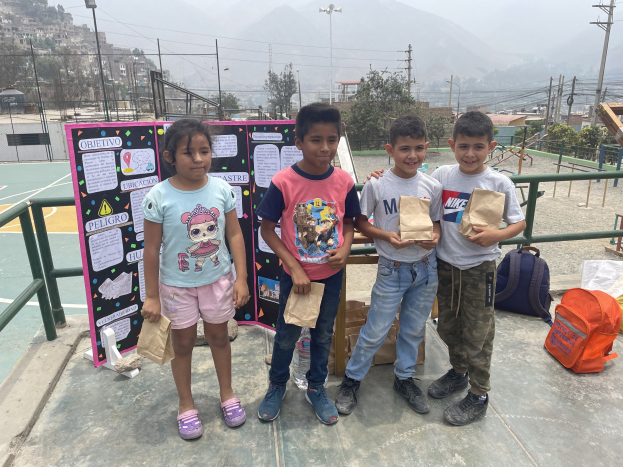 Gruppe von Kindern auf einem Basketballfeld, die Papiertüten halten, mit Tüten und Gegenständen rechts und einer Tafel mit Text links, vor dem Hintergrund von Zäunen, Pfosten, Straßenlaternen, Bäumen, Gebäuden, Hügeln und Himmel.