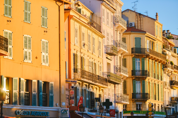 Eine Stadtstraße mit hohen Gebäuden, die Fenster, Geländer, Balkone und verschiedene Straßenausstattungen unter einem klaren blauen Himmel zeigen.