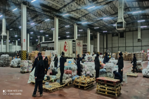 Eine Gruppe von Menschen, die um ein großes Lagerhaus mit Säcken voller Lebensmittel, Holzpaletten, Containern, Pfählen, einem Schild, einer Wand, einem Dach mit Deckenlampen und Metallpfählen sowie einem Wasserzeichen auf der linken Seite herumstehen, das den Anschein eines Lebensmittelverteilungszentrums in Dubai erweckt.