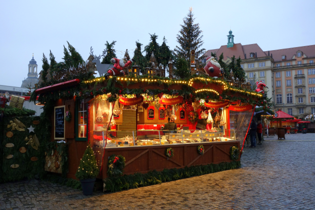 Ein Weihnachtsmarktstand auf einem Kopfsteinpflasterweg, umgeben von Gebäuden, beleuchteten Bäumen und Menschen, mit einem sichtbaren Himmel im Hintergrund.