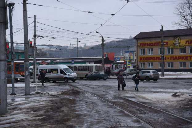 Gruppe von Menschen, die neben einer Bahnschiene auf einer Straße mit Fahrzeugen, Strommasten, Ampeln, Gebäuden, Bäumen und einem Lieferwagen gehen.