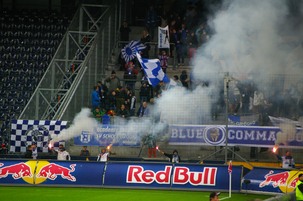 Eine Gruppe von Menschen steht auf einem Fußballfeld innerhalb eines Stadions, einige halten Fahnen und Schilder, mit Rauch, der vom Boden aufsteigt und leeren Stühlen auf der linken Seite sichtbar.