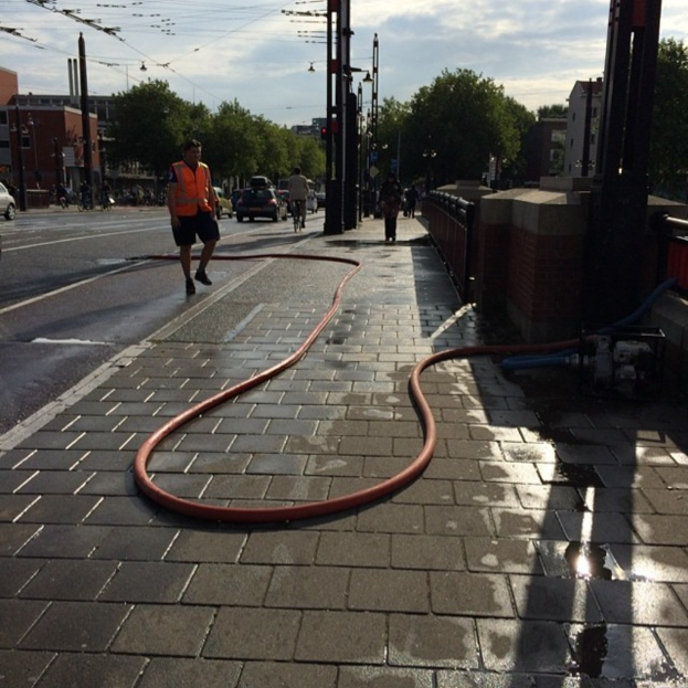 Eine Straße mit fahrenden Fahrzeugen auf der linken Seite, ein Gehweg mit einem Wasserrohr und einem Geländer auf der rechten Seite, eine Wand hinter dem Geländer und Bäume, Gebäude und einen Pfahl im Hintergrund unter einem Himmel.