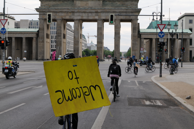 Eine Gruppe von Radfahrern fährt am Brandenburger Tor in Berlin vorbei, einer hält eine gelbe Tafel, mit Laternen, Ampeln, Gebäuden, Bäumen und einem klaren Himmel im Hintergrund.