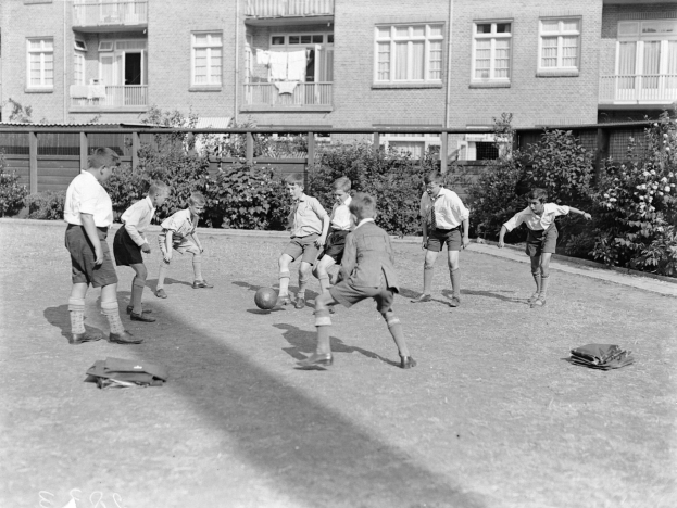 Schwarzes Foto von jungen Jungs, die auf einem Rasenfeld Fußball spielen, mit einem Ball in der Mitte, Gebäuden mit Fenstern, Geländern und Balkonen im Hintergrund und Pflanzen und Bäumen drumherum.