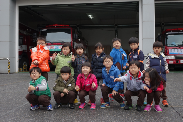 Kinder posieren vor einer Feuerwache, mit sichtbaren Feuerwehrautos in der Garage und einem Geländer auf der linken Seite.