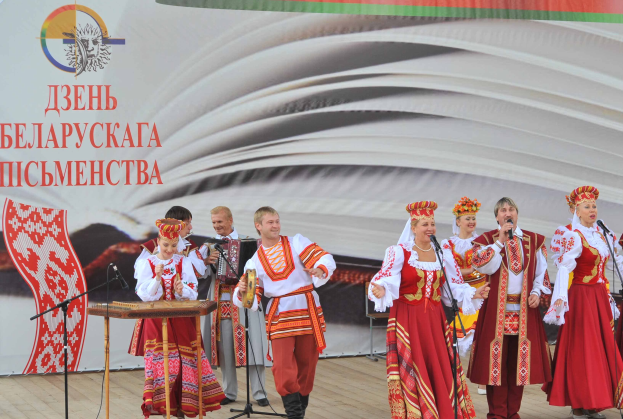 Eine Gruppe von Menschen in roter und weißer Kleidung auf einer Bühne, die singen und Musikinstrumente spielen, mit Mikrofonen und einem Banner mit der Aufschrift "Ukrainischer Folklore-Festival" im Hintergrund.