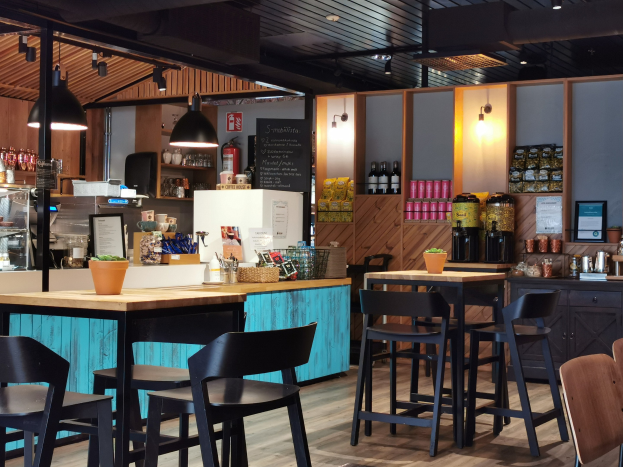 Innenraum eines Restaurants mit Holztischen und -stühlen, einem Blumentopf, einer Tafel, einem Kühlschrank, einem Feuerlöscher, Regalen mit Flaschen, Schränken mit Gegenständen, einem Rahmen mit einem Foto an der Wand und Deckenleuchten.