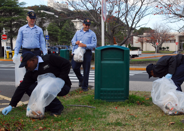 Drei Polizisten in Uniform sammeln Müll an einer Straßenrand mit Bäumen, Gebäuden und Fahrzeugen im Hintergrund.