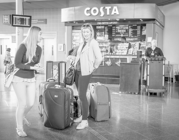 Zwei Frauen mit Gepäcktaschen vor einem Kaffeehaus an einem Flughafen stehend.
