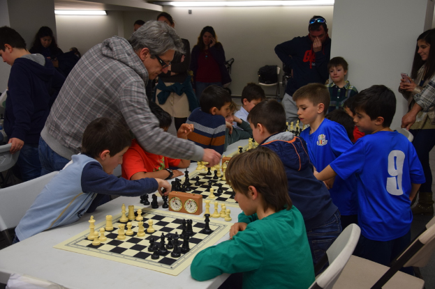 Kinder spielen Schach bei einem Wettbewerb, während Erwachsene im Hintergrund und an den Seiten zuschauen.