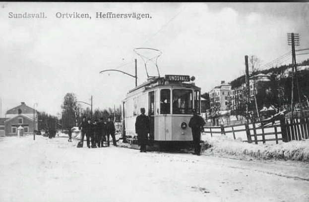 Ein Schwarz-Weiß-Foto einer Tram auf einer verschneiten Straße mit Menschen daneben, ein Zaun auf der rechten Seite und Gebäude, Bäume, Pfosten und Drähte im Hintergrund.
