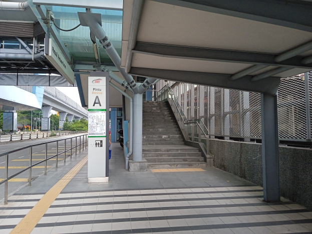 Ein Bahnsteig mit Treppe, eine Straße mit Geländer auf der linken Seite und ein Hintergrund mit Bäumen, Pfählen, Gebäuden und einer Brücke, mit Rohren und einer Tafel oben auf dem Bahnsteig.