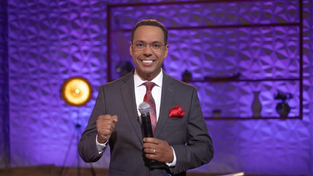 Stephen Colbert, Moderator von *The Daily Show with Trevor Noah*, steht auf einer Bühne und hält ein Mikrofon, trägt einen Anzug, Krawatte und Brille, wobei ein Scheinwerfer und ein Ständer hinter ihm sichtbar sind.