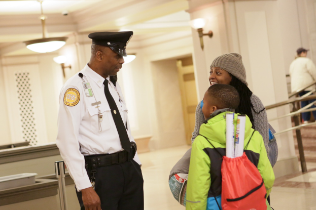 Ein Polizeibeamter spricht mit einer Frau mit Mütze und einem Kind an einem Flughafen, mit Menschen und wandmontierten Lichtern im Hintergrund.