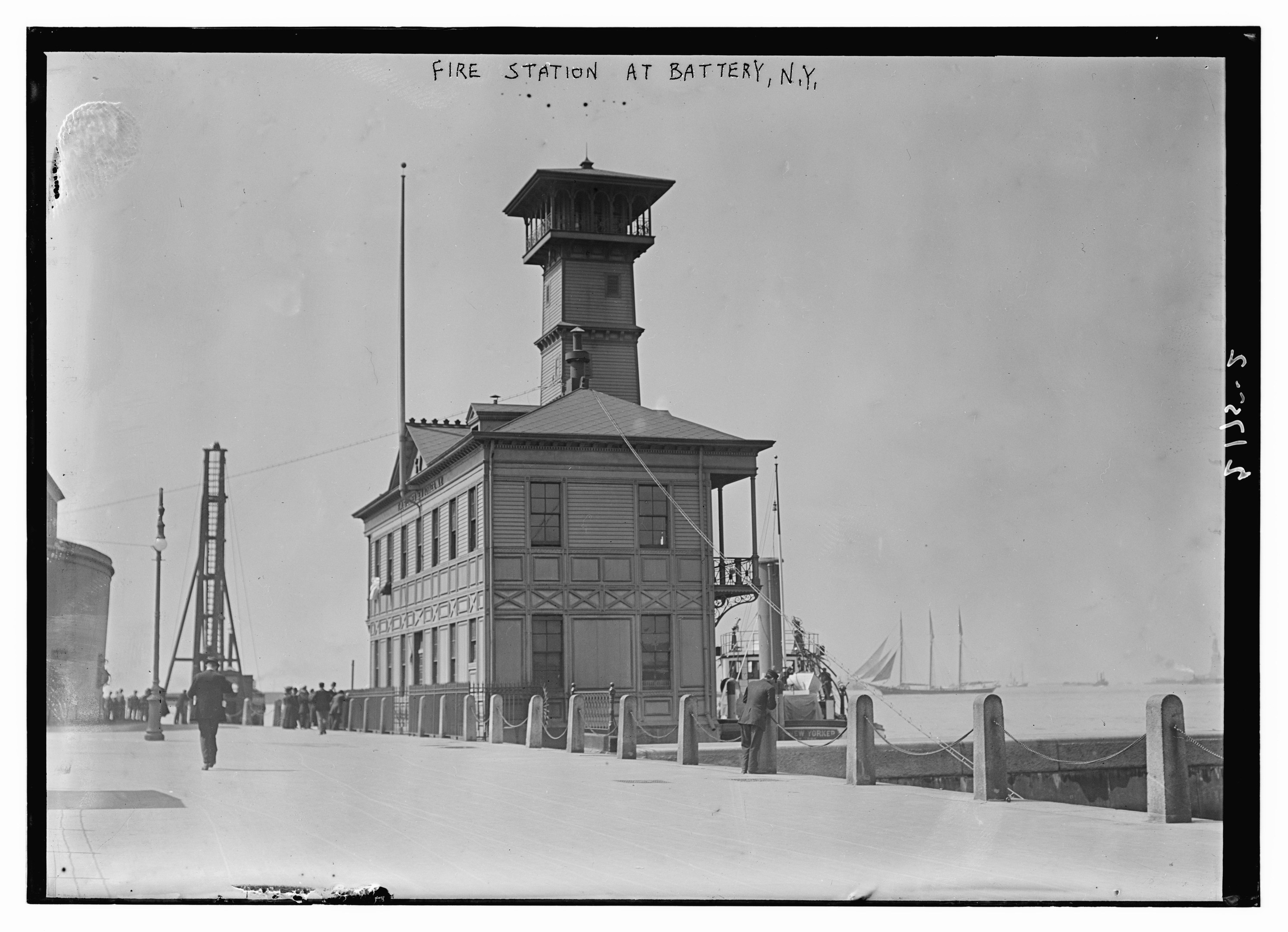 Schwarz-weiß-Foto einer Feuerwache in Battery, New York, mit einem Gebäude, Menschen, Pfählen, einem Zaun, Booten auf dem Wasser und einem bewölkten Himmel.