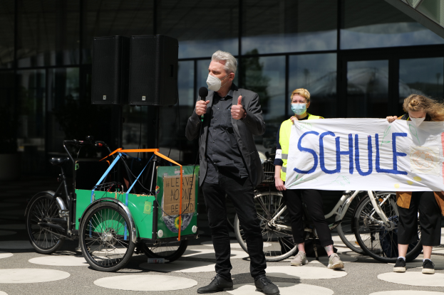 Ein Mann mit einer Maske hält ein Mikrofon vor eine Gruppe von Menschen, einige halten ein Transparent mit Text, Fahrräder, ein Fahrzeug mit einer Kiste und Lautsprecher, sowie ein gläsernes Gebäude im Hintergrund.