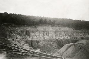 Ein Schwarz-Weiß-Foto einer Baustelle mit einer Brücke im Hintergrund, umgeben von Bäumen und einem klaren Himmel, mit Eisenbahnschienen und Steinen am unteren Bildrand.