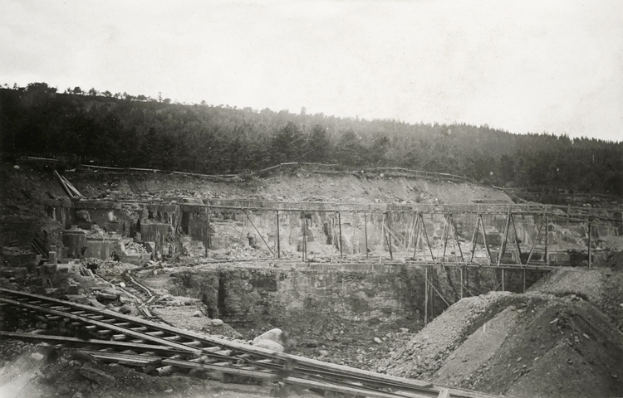 Ein Schwarz-Weiß-Foto einer Baustelle mit einer Brücke im Hintergrund, umgeben von Bäumen und einem klaren Himmel, mit Eisenbahnschienen und Steinen am unteren Bildrand.