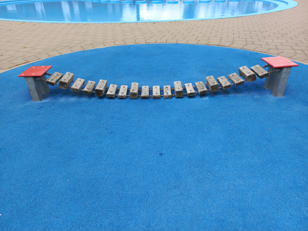 Ein Spielplatz mit blauer Oberfläche und einer roten Bank in der Mitte, umgeben von einem Schwimmbad mit Säulen im Hintergrund und einem Weg am Boden.