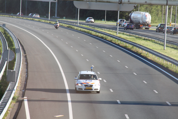 Ein Auto auf einer Autobahn mit einem stationären Polizeiwagen am Straßenrand, andere Fahrzeuge, Leitplanken, Strommasten, Schilder und Bäume im Hintergrund.