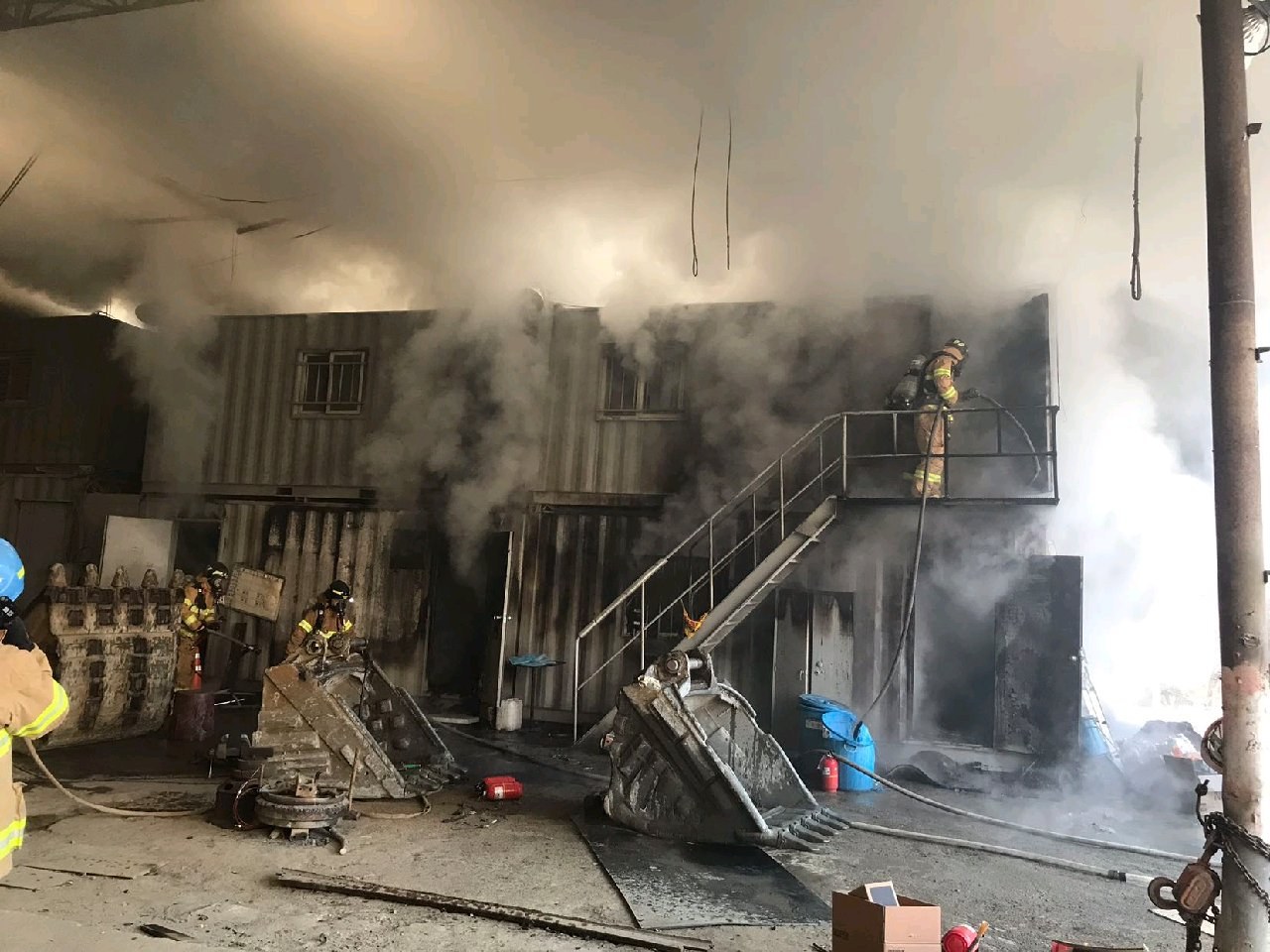 Feuerwehrleute in Helmen arbeiten daran, ein Lagerfeuer zu löschen, mit Rauch, der aus dem Gebäude aufsteigt, Treppenstufen, die zur Tür führen, eine Kette an einem Pfahl, verstreute Trümmer auf dem Boden und Fenster im Hintergrund.