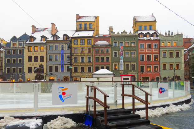 Eisbahn in einem Stadtplatz umgeben von Gebäuden, Treppen, Geländern, Informationsschildern, schneebedecktem Boden, Laternen, Bäumen und einem Himmel im Hintergrund.