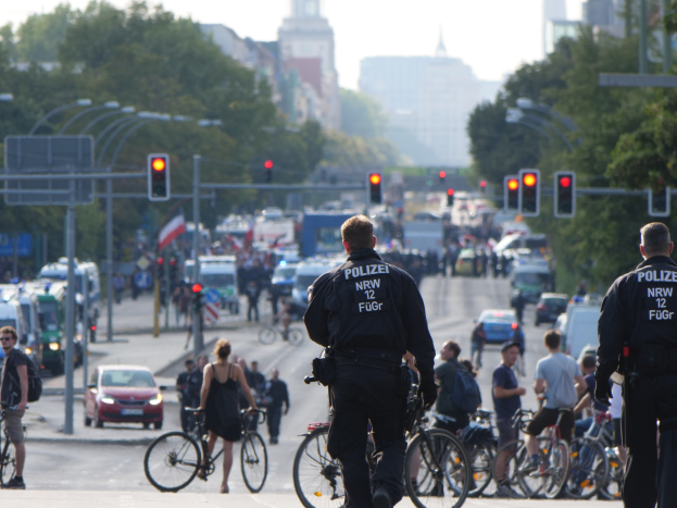 Zwei Polizeibeamte stehen neben einer Gruppe von Menschen auf Fahrrädern an der Straße, mit Fahrzeugen, Verkehrszeichen, Bäumen, Gebäuden und einem klaren blauen Himmel im Hintergrund.