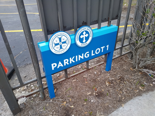 Blau-weißes "Parkplatz 1"-Schild vor einem Metallzaun, mit einer Straße und einem Baum im Hintergrund.
