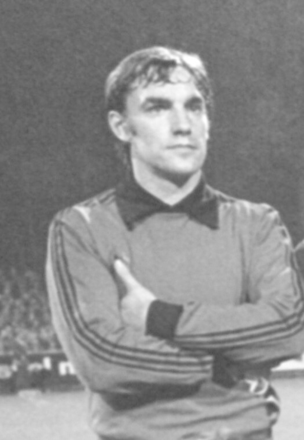 Ein Schwarz-Weiß-Foto eines Mannes, der mit verschränkten Armen auf einem Fußballfeld steht und von einer Gruppe von Menschen im Hintergrund umgeben ist, die angeblich der legendäre Torwart von Manchester United ist.