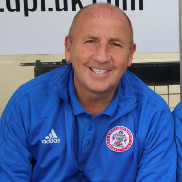 Zwei Männer in einem Raum, einer in einer blauen Jacke, der neben einem anderen Mann lächelt, mit einer Tafel an der Wand dahinter.