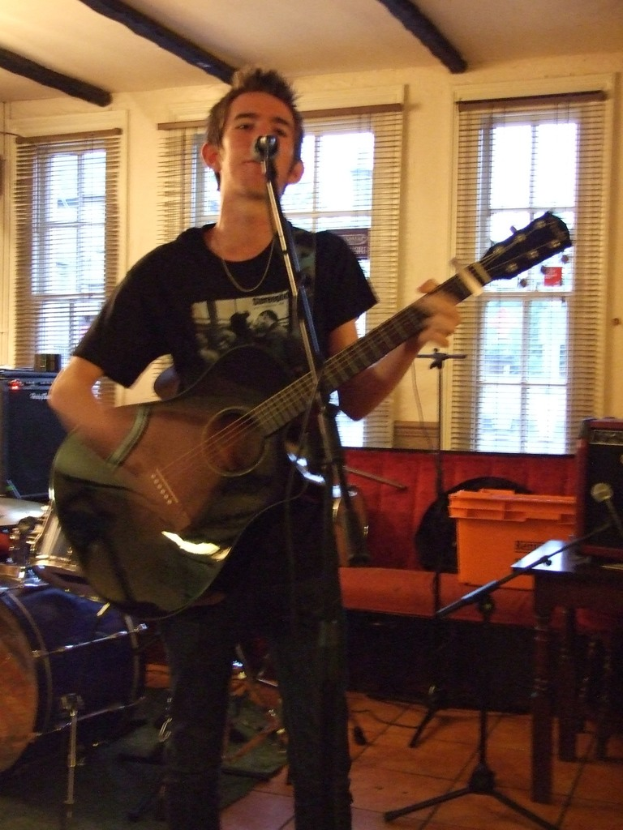 Ein Mann in schwarzem T-Shirt und Hose steht mit einer Gitarre und einem Mikrofon auf einem Ständer, mit einer blauen Trommel in der linken Ecke, einem Sofa mit Gegenständen darauf und durchsichtigen Vorhängen im Hintergrund unter einer Decke.