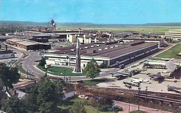 Altes Flughafen-Foto zeigt geparkte Flugzeuge, Gebäude, Bäume, Straßen, Fahrzeuge, Pfosten, Gras, Bahnschienen, Türme, Hügel und einen klaren blauen Himmel mit Text unten.