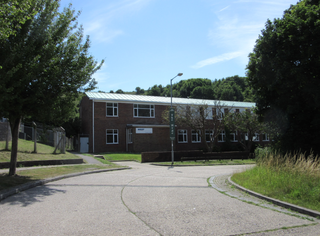 Ein Schulgebäude mit Fenstern, einem Straßenpfahl, einem Schild, einer Baumgruppe, Gras, einem Zaun und einem bewölkten Himmel, das sich in einem Park mit saftig grünem Gras befindet.