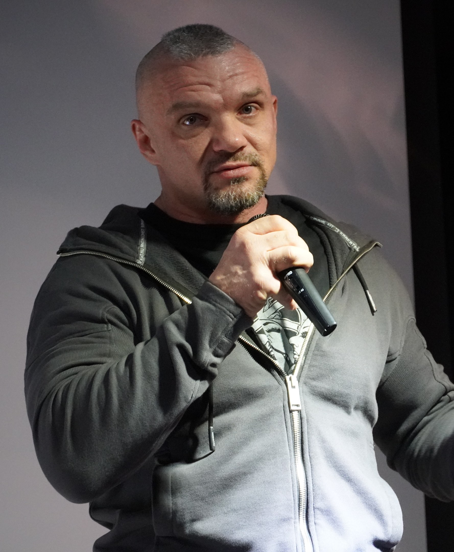 Ein Mann in einer schwarzen Kapuzenjacke und einem schwarzen Anzug mit einem weißen Hemd, der ein Mikrofon hält, steht selbstbewusst vor einer Wand.