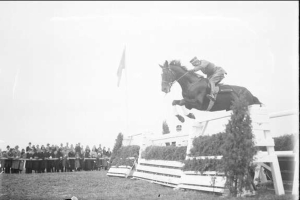 Schwarze und weiße Fotografie eines Pferdes und Reiters, die über ein Hindernis springen, bei den Royal Ascot Horse Trials 1953, mit Zuschauern links, einer Flagge im Hintergrund und Gras unten.