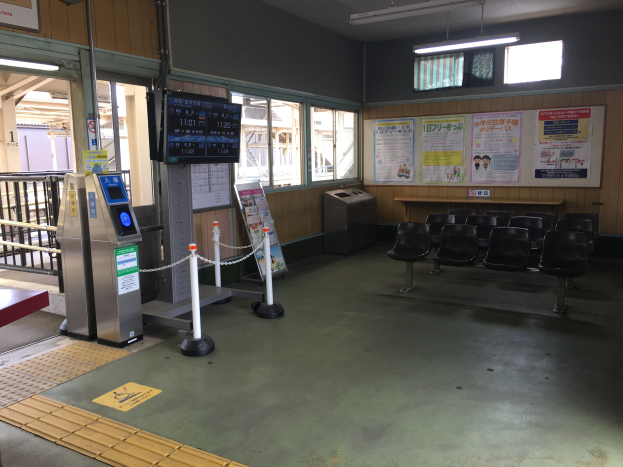 Ein belebter Bahnhofseingang mit Reihen von Stühlen, einem Service-Tresen, Ticketautomaten, Deckenleuchten, Geländern, Informationsanzeigen und Wandplakaten.