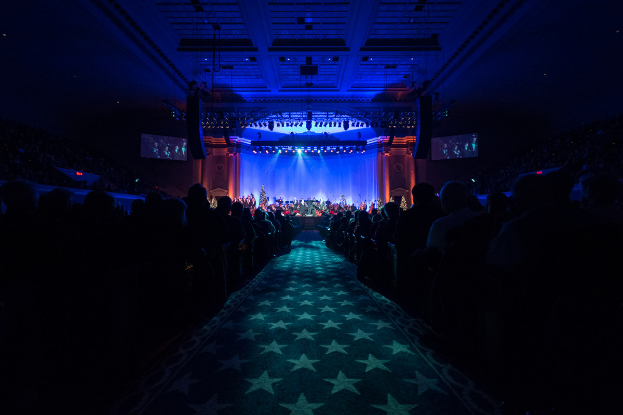 Ein großer Zuschauerraum mit Sitzplätzen, eine Bühne mit Musikern und eine Decke mit Lichtern und Fernsehern, was auf einen Konzertsaal für die Nationale Convention 2019 hindeutet.