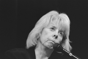 Ein Schwarz-Weiß-Foto einer Frau, die in ein Mikrofon singt und ein weißes Hemd trägt, mit zurückgebundenem Haar, geschlossenen Augen und zusammengelegten Händen vor einem unscharfen, verträumten Hintergrund.