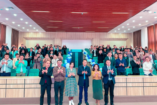 Eine Gruppe von Menschen steht vor einem Publikum, einige klatschen, in einem Raum mit Stühlen, Tischen, Luftballons, Vorhängen und Deckenlampen, bei der Eröffnungsfeier einer neuen Schule.