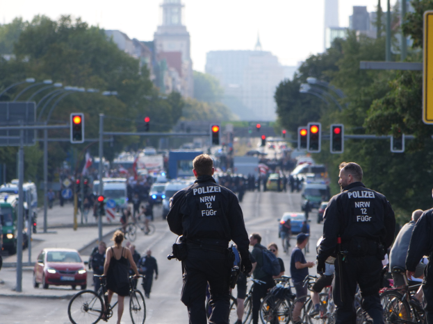 Polizeibeamte fahren mit Fahrrädern eine straßenbepflanzte Straße entlang mit Gebäuden und einem klaren blauen Himmel im Hintergrund.