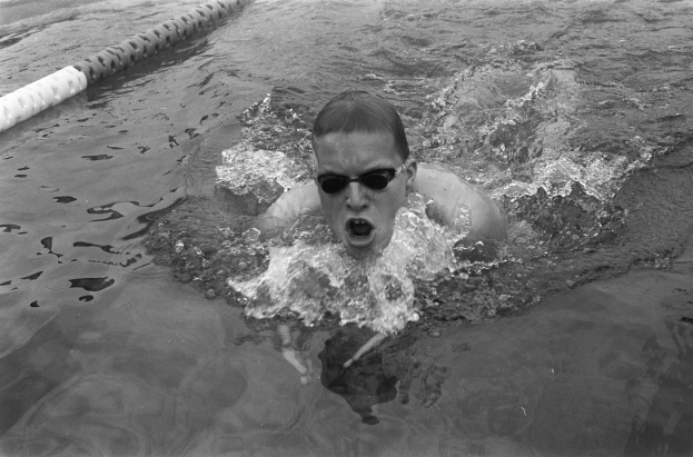 Ein Mann schwimmt in einem Pool während eines Rennens, trägt eine Schwimmbrille und ist von Bahntrennern umgeben, dargestellt in Schwarz-Weiß.